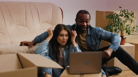 The mixed couple is sitting on the floor next to the couch, watching a sports Stock Footage 235731100