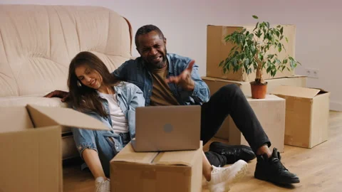The mixed couple is sitting on the floor next to the couch watching a sports Video stock 235773747