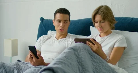 Mixed race couple is lying in bed, browsing in smartphone and tablet computer Stock Footage 114918598