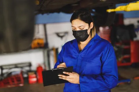 Mixed race female car mechanic wearing face mask and overalls, making notes on Stock Photos