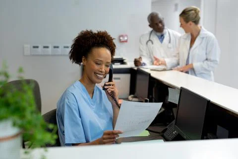 Mixed race female doctor at desk in hospital reception talking on phone and Stock Photos