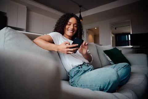 Mixed race female smiling while relaxing on couch typing on cellular device High Stock Photos