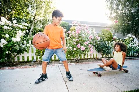 Mixed Race girl on skateboard watching brother dribbling basketball Stock Photos