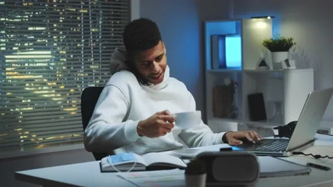 Mixed-race man working on computer, speaking by smartphone and drinking a cup of Stock Footage 131537697
