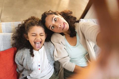 Mixed race mother and daughter sitting on sofa doing funny faces, taking selfie Stock Photos