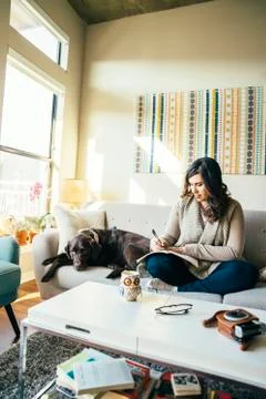 Mixed Race woman on sofa with dog writing in journal Stock Photos