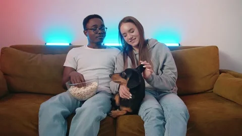 A mixed young couple is sitting on the couch having fun playing with their Stock Footage 173015717