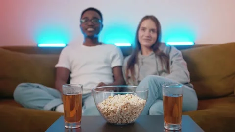 Mixed young couple watching fun movie while sitting on sofa at table with drinks 库存影片 173413445