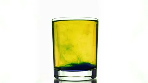 Mixing using glass stick of blue pigment dissolve at yellow colored water glass Stock Footage 154438120