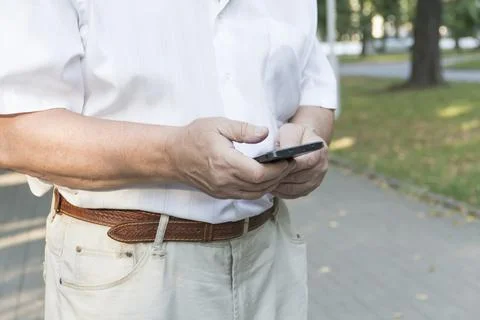 A mobile phone in the large hands of an elderly man in a white shirt on a wal Stock Photos