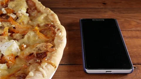 The mobile phone rests on a wooden table next to a homemade pizza with different Stock Footage