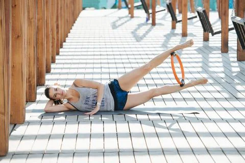 Model doing side hollow hold with a pilates hoop outdoors under a wooden frame Stock Photos