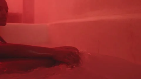 Model lying in bath rubs leg and looking straight at camera in red light Stock Footage 275352288