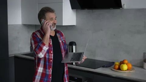 Modern aged man using laptop in kitchen and talking by phone at the same time Stock Footage 201475124