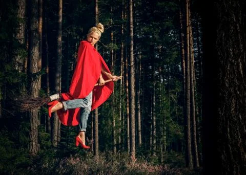Modern angry witch in jeans and red cape flying on a broom through the forest Stock Photos
