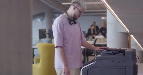A modern, blond-haired man efficiently operates a high-tech paper printer in a Stock Footage 254524399