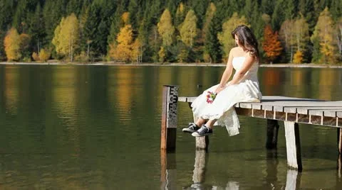 Modern bride with sneakers, holding flowers bouquet sitting on lake dock Stock Footage