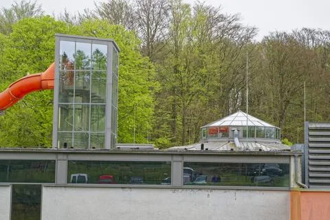 Modern building with glass elevator and orange slide surrounded by green tree Stock Photos