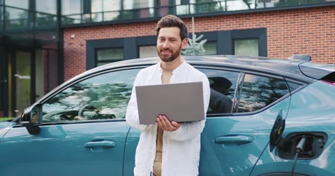 Modern businessman working on laptop while his electric car charges at a city st Stock Footage 319309992