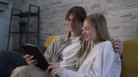 Modern couple is sitting on the couch, looking at a computer tablet and talking Video stock 259329801
