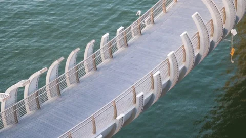 A modern curved pedestrian bridge spans over tranquil blue water Vídeos de archivo 336400780