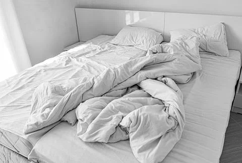 A modern double bed features messy white sheets, a crumpled duvet, and soft Stock Photos