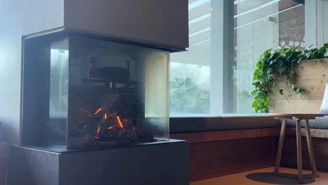 Modern fireplace burning in cozy interior with large windows and green plants Stock Footage 313685336