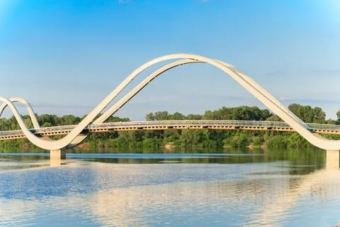 Modern infinity loop bridge gracefully arches over a calm river with lush g.. Stock Photos