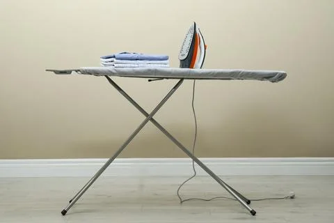 Modern iron and stack of clean clothes on board against beige background 스톡 사진