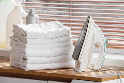 Modern iron with stack of clean towels on commode near window in bathroom Stock Photos