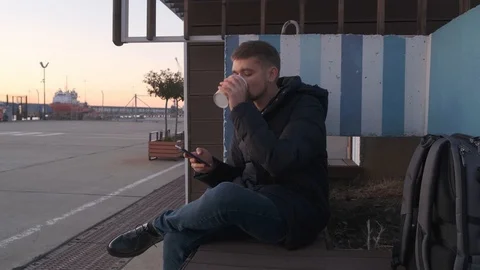 Modern man in casual clothes, sitting on a bench drinking coffee and using the Stock Footage 123209331
