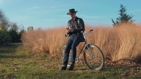 Modern man in a hat, standing in the Park leaning on a Bicycle and uses the Video stock 121807605