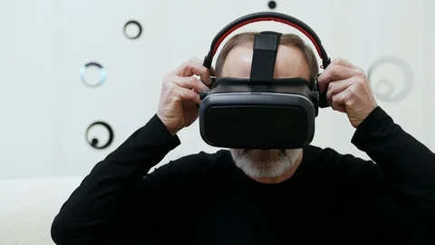 Modern middle-aged man with grey beard puts on wireless headphones and use VR Stock Footage 124618825