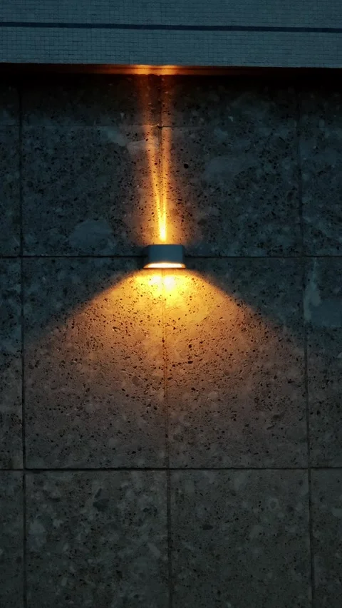 Modern outdoor wall lamp casting warm yellow light on grey stone facade Stock Footage 334632910