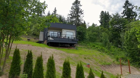 Modern rectangular cabin on a steep green hill surrounded by forest and small Stock Footage 320393336