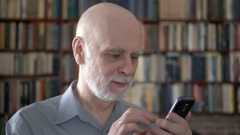 Modern senior man at home using cellphone, browsing, reading. Bookcase Stock Footage 74756477