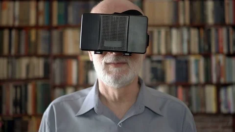 Modern senior man at home using virtual reality glasses. Bookcase bookshelves in Stock Footage 75334714