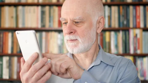 Modern senior man at home using cellphone, browsing, reading. Bookcase Stock Footage 86327382