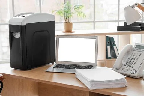 Modern shredder with documents and blank laptop on desk in office Stock Photos