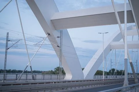 Modern white arch bridge spanning water, railway tracks, overhead power lines Stock Photos