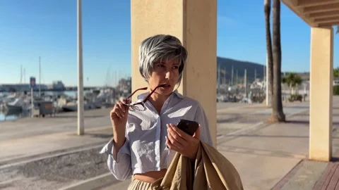 Modern woman over 50 checking her phone by a harbor pergola Stock Footage 346293960