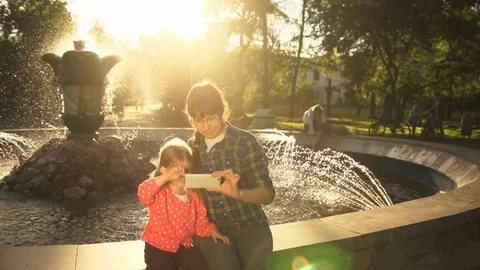 Mom and daughter take selfi on the phone at the fountain in the summer at sunset Stock Footage