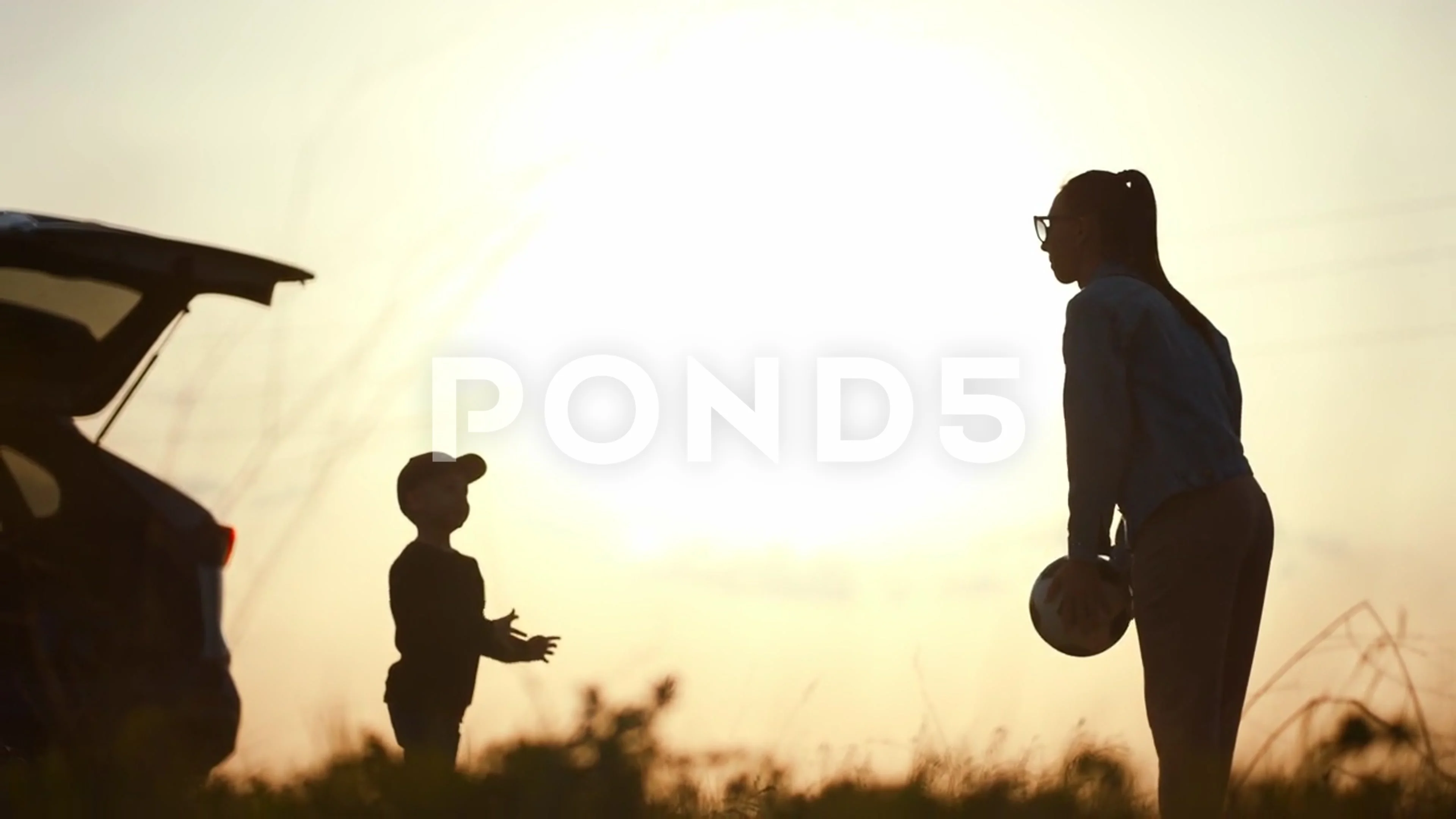 Mom and son play on ball near a car with an open trunk at sunset