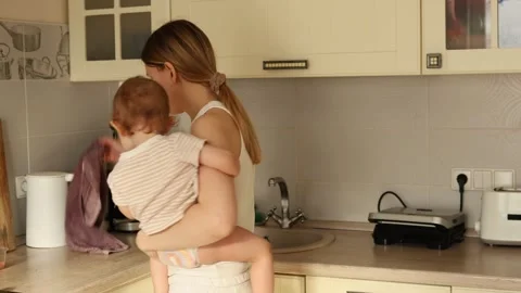 Mom cooks in kitchen while holding baby during morning routine Stock Footage 297849011