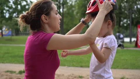 Mom helps his son put on a helmet to safely ride a bike in the city park Stock Footage 78480603