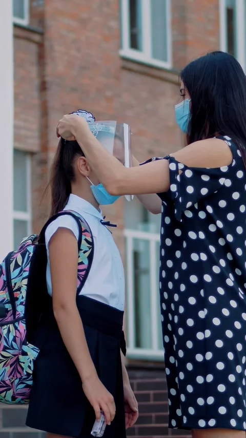 Mom puts protective face shield on her daughter schoolgirl, on school building Vídeos de archivo 316778840