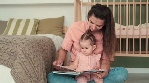 Mom reads a book to the kid while sitting near the bed at home Stock Footage 156472802