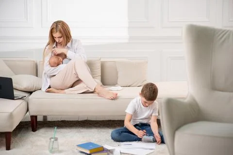 Mom sits on the couch and feeds the baby. The son has coloring pages on the Stock Photos