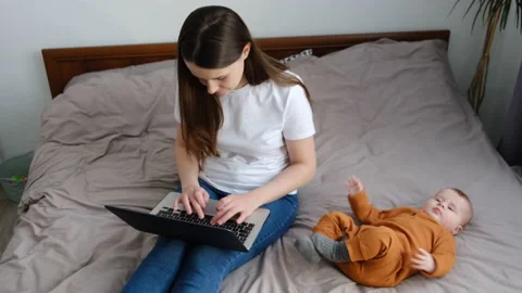 Mom using laptop notebook looking at screen typing message sit on bed near baby Stock Footage 151084088