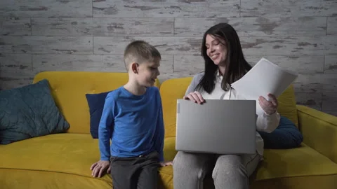 Mom is working on the couch with documents and a laptop, her son comes up to her Stock Footage 259681836